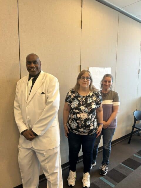 African American man stands against a white wall in a white suit, next to a blond woman with glasses, black pants and a flower top, next another woman with a pony tail, jeans and a gray shirt with a white stipre
